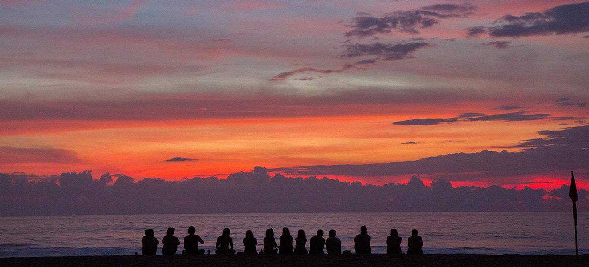 Silhouettes of a group of people sitting on the beach, watching a vibrant sunset with shades of red, orange, and purple over the ocean.