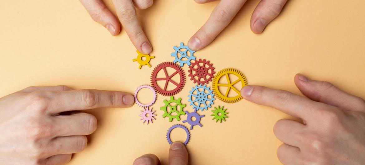 Hands pointing at interconnected colorful gear pieces on a neutral background, symbolizing teamwork, collaboration, and system integration