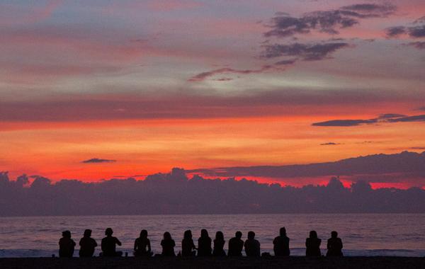 Silhouettes of a group of people sitting on the beach, watching a vibrant sunset with shades of red, orange, and purple over the ocean.