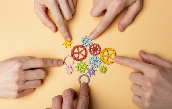 Hands pointing at interconnected colorful gear pieces on a neutral background, symbolizing teamwork, collaboration, and system integration